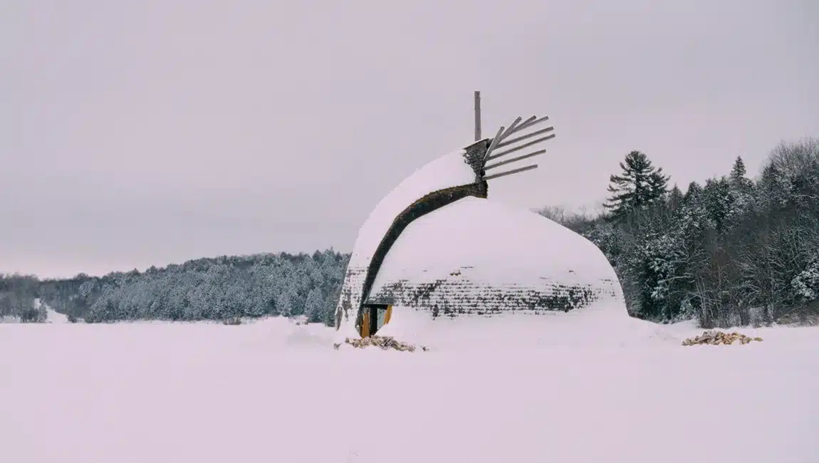 Still from AKI, directed and produced by National Screen Institute alum Darlene Naponse. A snow-covered dome-shaped building stands alone in a vast white field during winter. The structure features a curved, sculptural design with dark wood siding partially visible beneath the snow and a distinctive fan-like element extending from the roof. A line of snow-covered trees stretches across the background beneath an overcast grey sky.