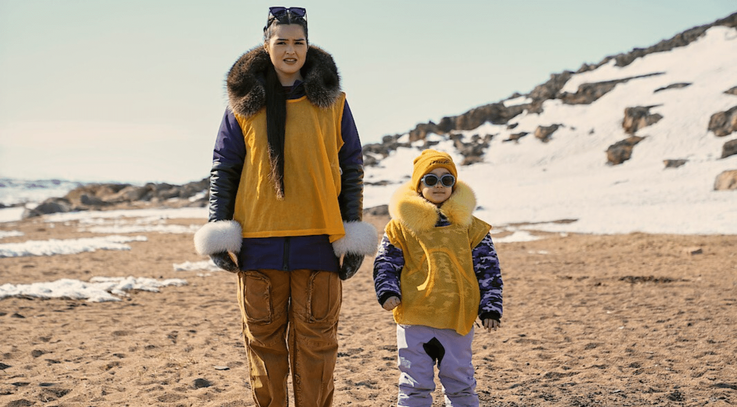 Two people stand on a sandy Arctic shoreline with patches of snow and rocky hills in the background. They are dressed in cold-weather clothing and matching yellow pinnies over purple outerwear. The adult wears fur-trimmed mitts and a fur-lined hood, while the child wears round sunglasses, a yellow toque and a fur-trimmed collar. Both face the camera under a clear sky in a remote northern landscape.