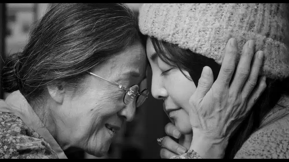 Black-and-white still of two people gently pressing their foreheads together. An older person wearing glasses cradles the younger person's face with both hands as they share a quiet, emotional moment with their eyes closed.