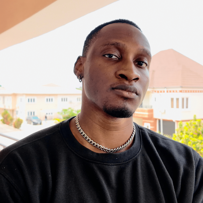 Alt text: A close-up portrait of a man wearing a black shirt, silver chain necklace and hoop earring, standing outdoors on a balcony with buildings in the background.