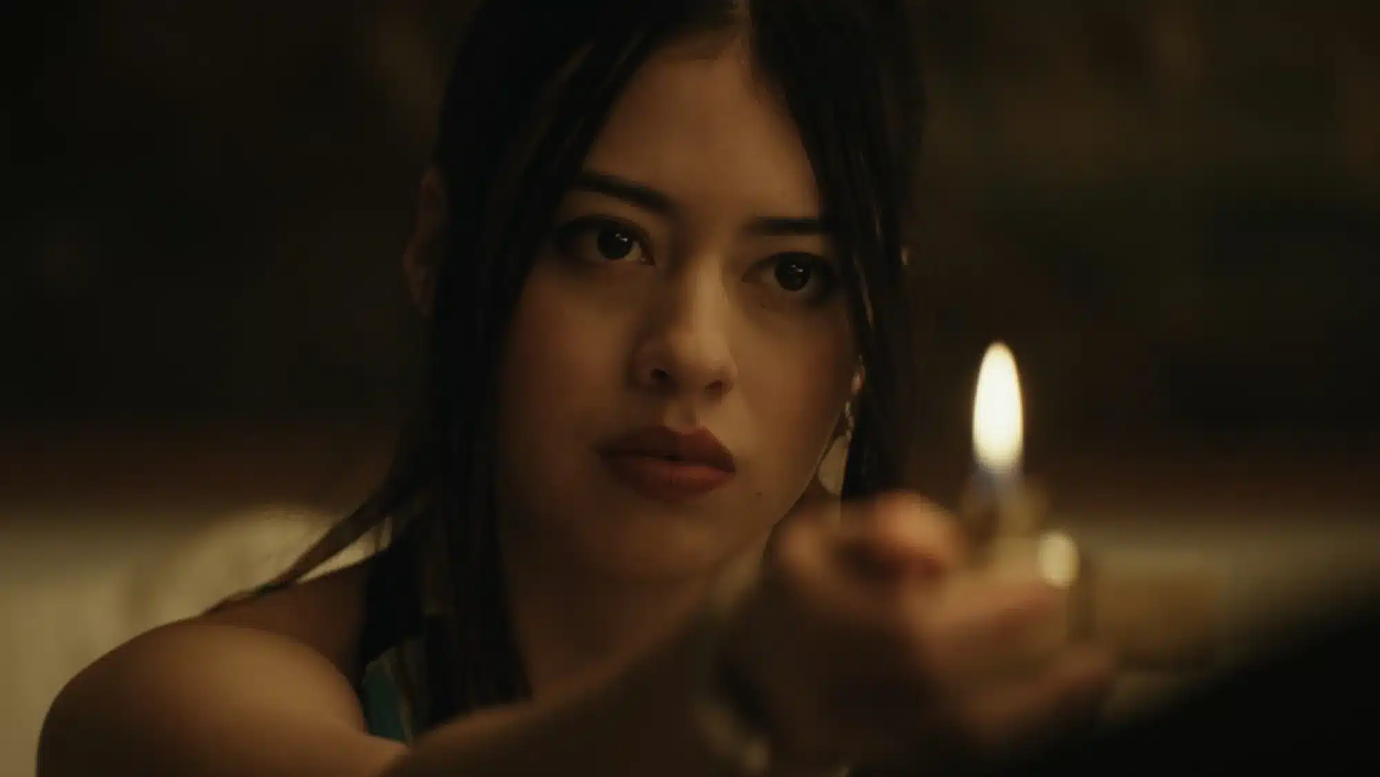 Close-up of a woman with dark hair holding a lit match in the foreground, her face softly illuminated against a dark, blurred background.