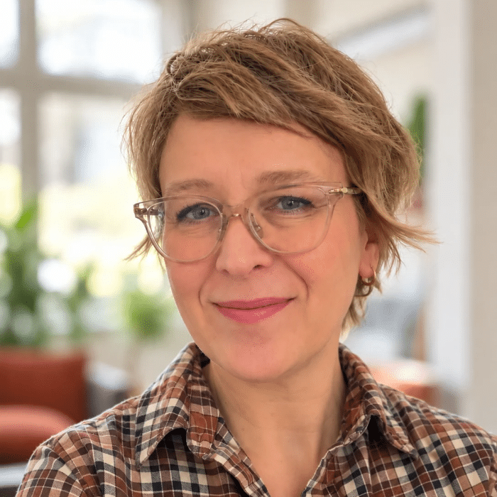 Headshot of Christine Kleckner smiling at the camera. She has short, light brown hair, clear-framed glasses and a brown plaid shirt. The background is softly blurred.