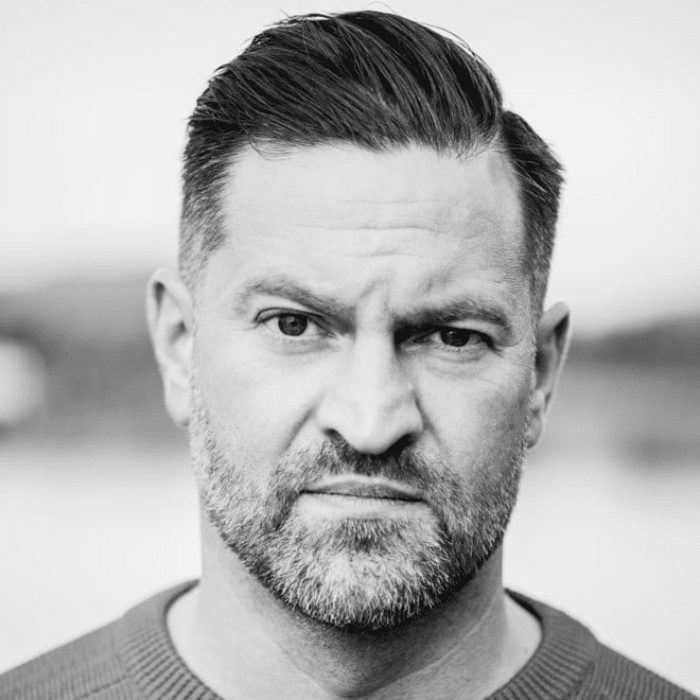 Black and white headshot of Adam Scorgie with short hair, beard and moustache wearing a dark t-shirt.
