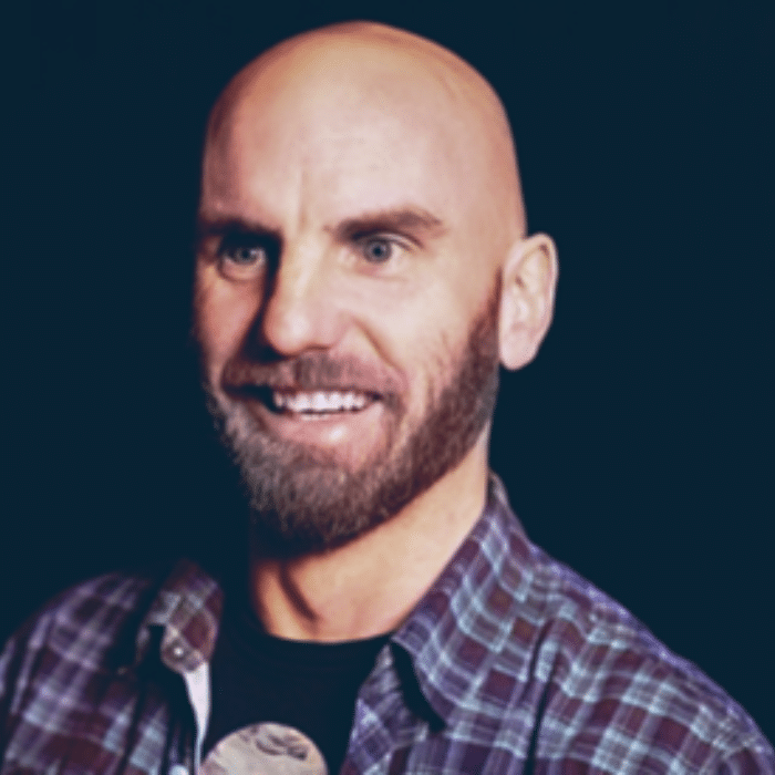 Cody Graham with a moustache and beard, wearing an open flannel shirt with a dark t-shirt underneath.