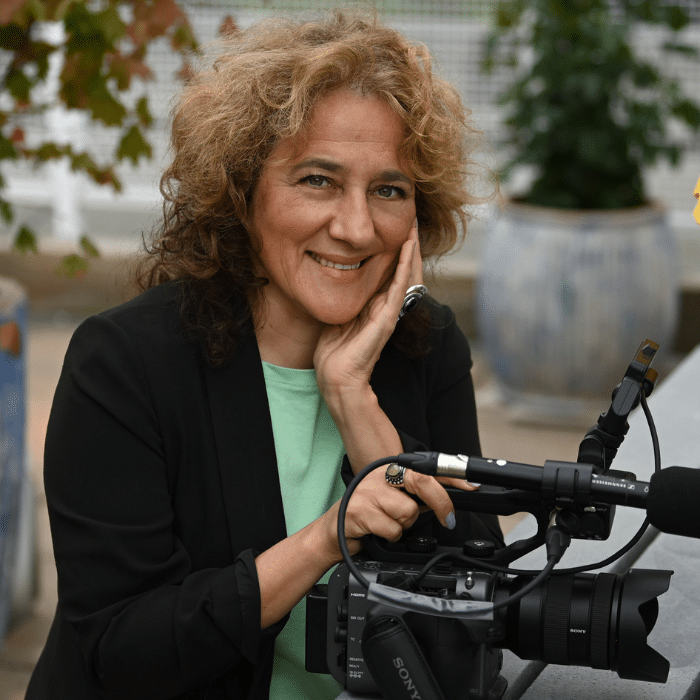 Julia Ivanova with curly brown hair wearing a black blazer with a light green shirt underneath, left hand resting on face, right hand resting on a video camera.