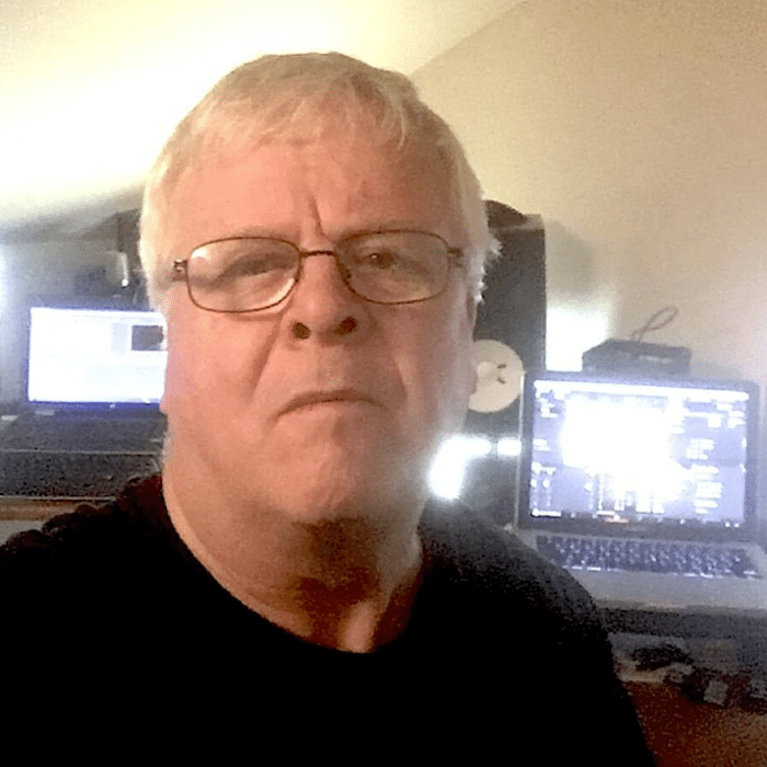 Mark Collings with short white hair, wearing glasses and a black shirt. In front of multiple computer screens.