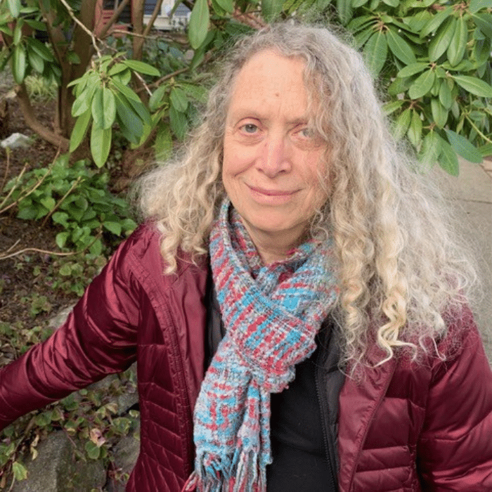 Peggy Thompson with long white blonde hair, wearing a plum jacket, a turquoise, red and white knitted scarf and a black shirt.
