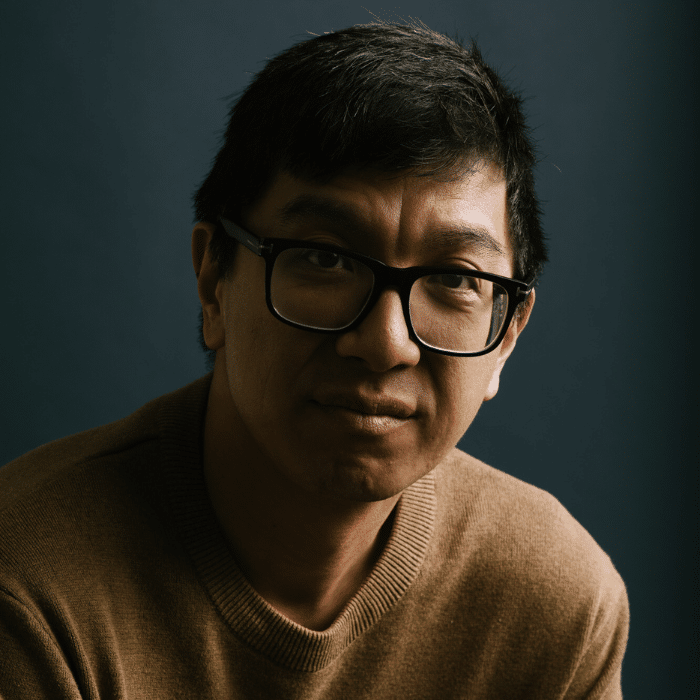 Ryan Mah partially in shadow with short black hair, wearing glasses and a light brown sweater.