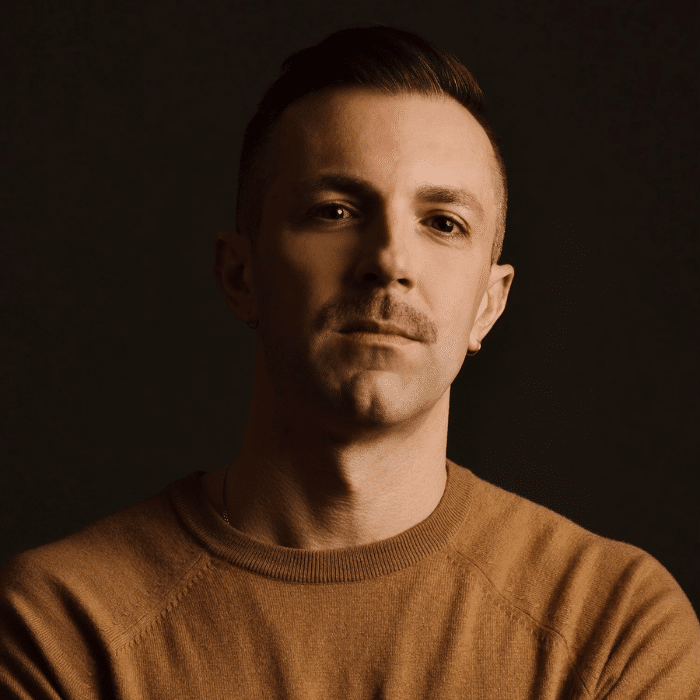 Sean Horlor partially in shadow with short facial hair wearing a brown shirt.