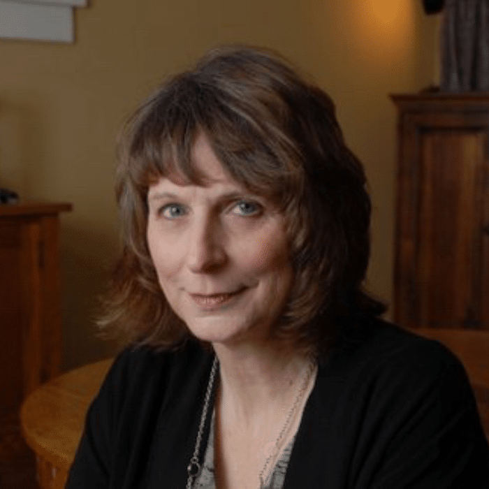 Sharon McGowan with brown hair, wearing a necklace, black cardigan and grey shirt.