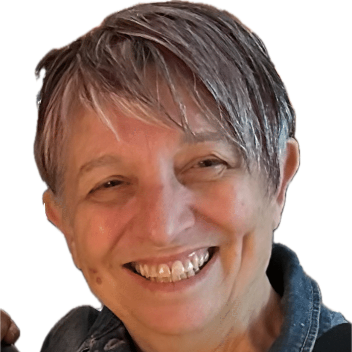 A person with short, gray-and-purple hair smiles warmly at the camera. She is wearing a denim jacket, and the background is plain white.