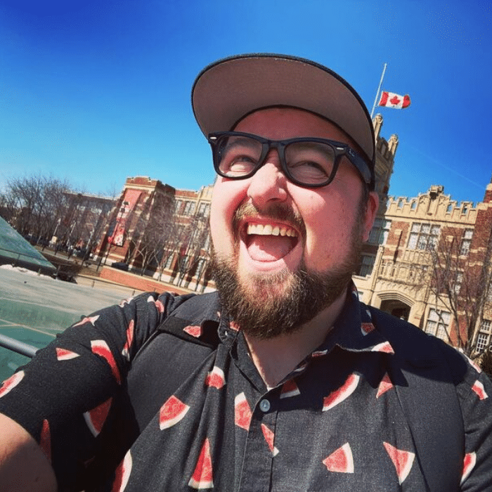 A person with a beard and moustache smiles outdoors on a sunny day. He is wearing black-framed glasses, a black shirt with a watermelon print and a baseball cap, with historic-looking brick buildings and a Canadian flag in the background.