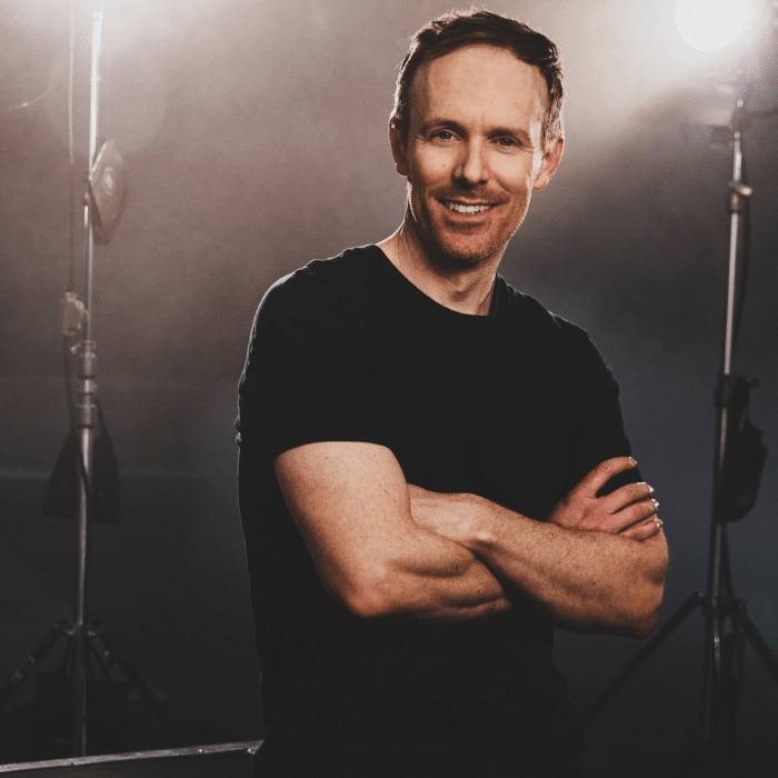 Kyle Rideout standing with arms crossed with short brown hair, dark t-shirt, arms crossed. Dark background with tall lights on both sides.