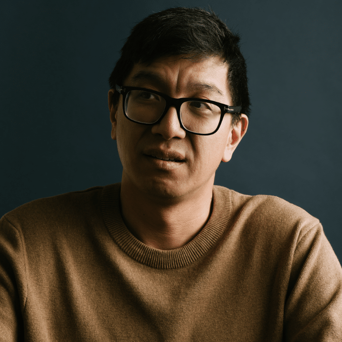 A person with short dark hair and black-framed glasses looking to the side. He is wearing a brown sweater, with a dark studio background.