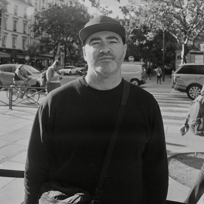 Black and white photo of Mike standing streetside wearing a dark-coloured baseball cap and a dark-coloured long-sleeved shirt and a crossbody bag.