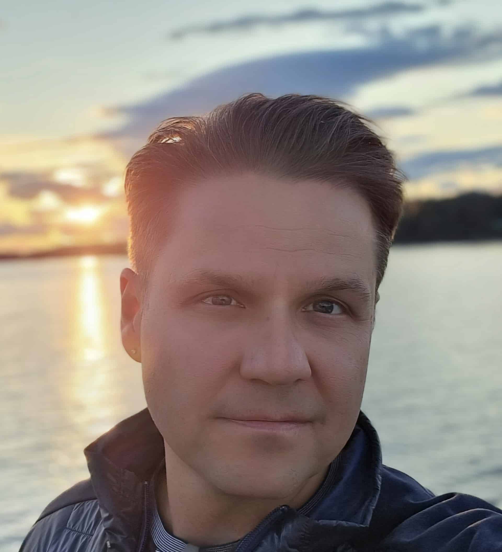 Jeff Stecyk with short brown hair, wearing long-sleeved shirt with collar up, standing in front of a a sunset over a body of water.