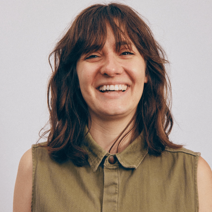 Sara Blake in front of a light grey background. She has shoulder-length reddish-brown hair with bangs. She is wearing an olive green, collared, button-up sleeveless shirt.