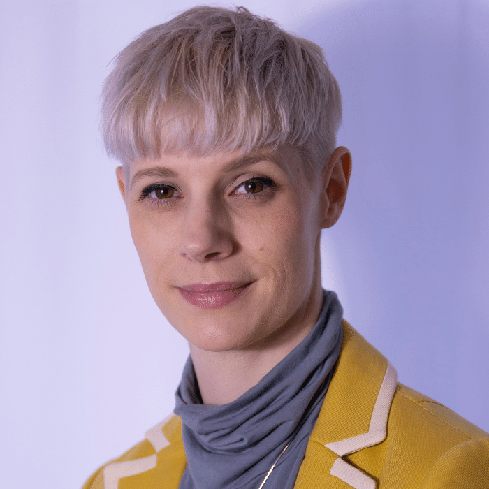 Lora Campbell in front of a light purple-grey background. They have short blonde hair and are wearing a grey turtleneck and a mustard yellow blazer with thick white piping.