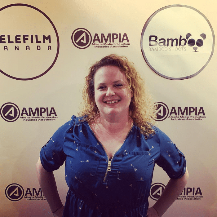 Sara Corry standing in front of a white background with black Telefilm Canada, AMPIA and Bamboo Shoots logos. She is wearing a blue short-sleeved dress with a zipper and an arrow pattern. Sara has curly, blonde shoulder-length hair.