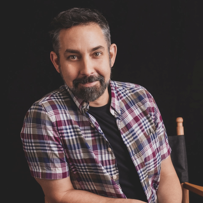 Kevin Eastwood seated on a director's chair in front of a black background. He has short dark brown and grey hair. He is wearing an unbuttoned plum, white, olive green and navy blue short-sleeved plaid shirt with a black t-shirt underneath.