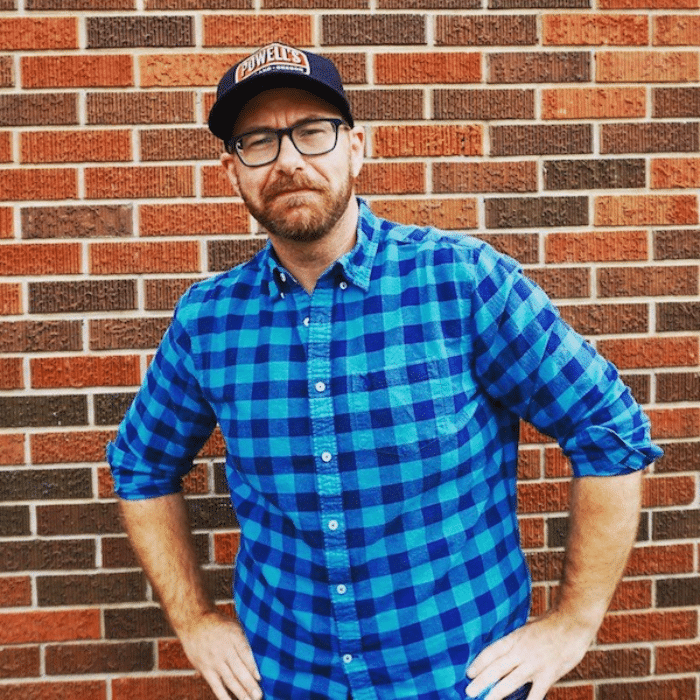 Jason Filiatrault standing with hands on hips in front of a brick wall. He is wearing a Powell's baseball cap, black glasses and a light and dark blue buttoned up plaid shirt. He has reddish-brown facial hair.