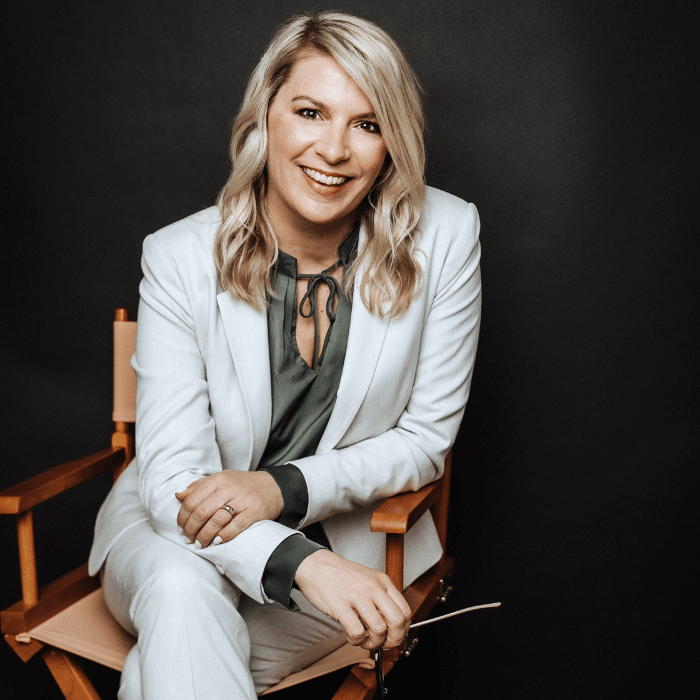 Kate Green is seated on a director's chair in front of a black background. She has wavy blonde hair and is wearing a light-coloured suit with a charcoal blouse.