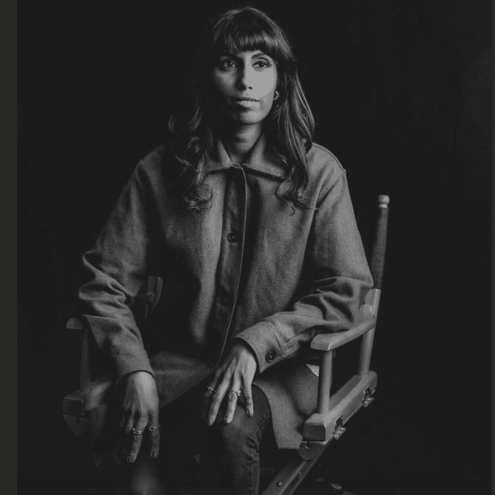 Black and white photo of Nimesha Mukerji seated on a director's chair. She has long wavy dark hair with bangs. She is wearing a button-up coat and dark jeans.