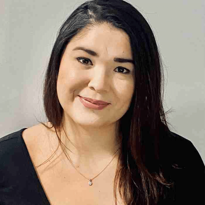 A woman with long dark hair, wearing a black top and a delicate pendant necklace. She is smiling softly and posed against a neutral background.