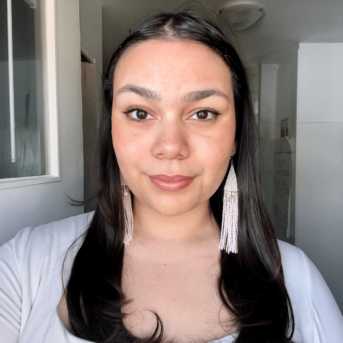 A woman with long dark hair and beaded tassel earrings, wearing a white top. She is looking directly at the camera, standing in front of a background with white walls and natural light.