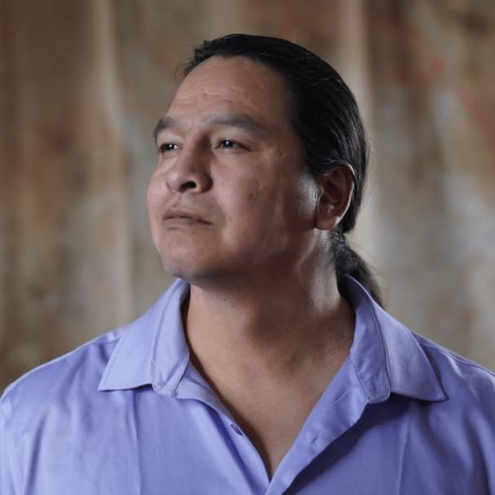 A man with long dark hair tied back, wearing a light purple collared shirt. He is looking thoughtfully into the distance, set against a softly blurred background.