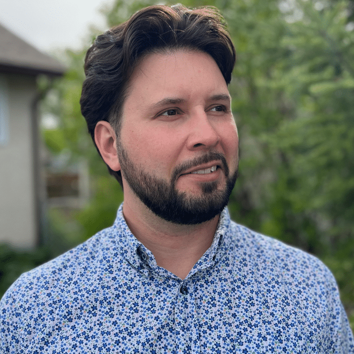 A man with short dark hair and a beard, wearing a blue and white floral button-up shirt. He is looking off to the side and is standing outdoors with greenery and a house in the background