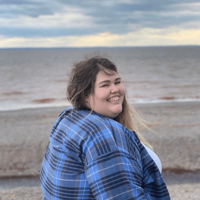 A woman with long hair smiles while looking over her shoulder, standing on a beach with waves and a cloudy sky in the background. She is wearing a blue plaid shirt.
