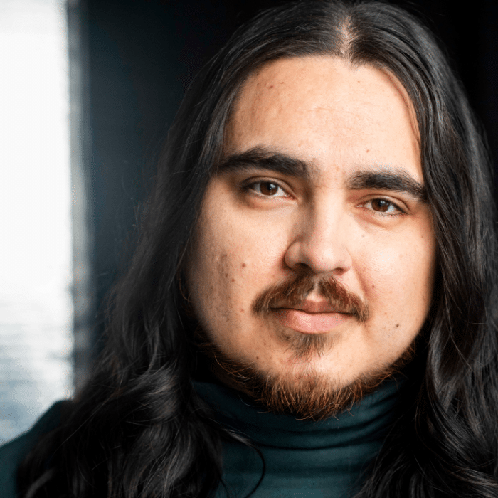 Close-up of a man with long dark hair and a beard, wearing a dark turtleneck. He is looking directly at the camera, against a softly lit dark background.
