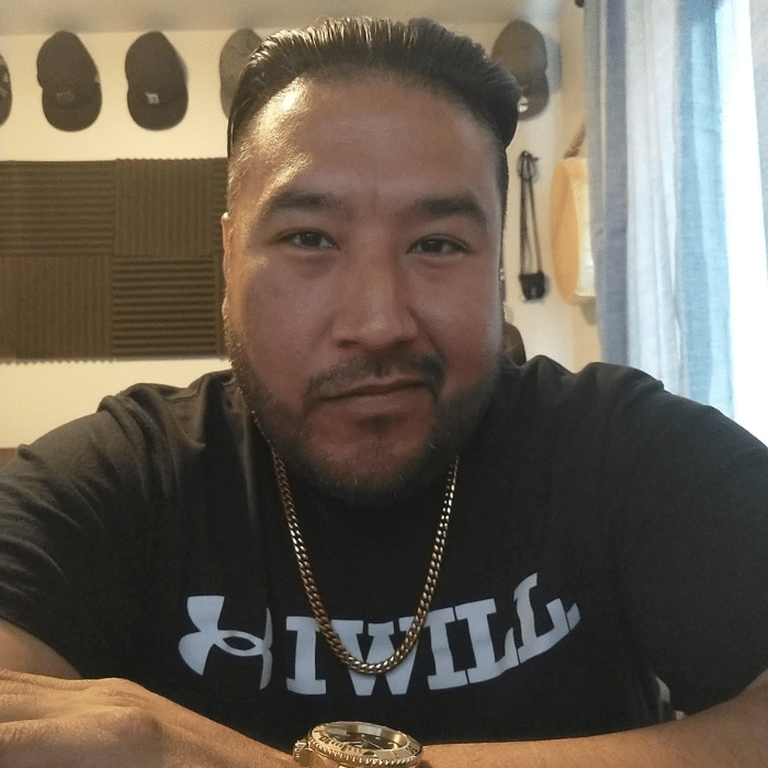 A man with slicked-back hair and a trimmed beard poses for a photo indoors. He is wearing a black Under Armour t-shirt that says 'I WILL,' a gold chain, and a gold watch, with soundproof panels and hats visible in the background.