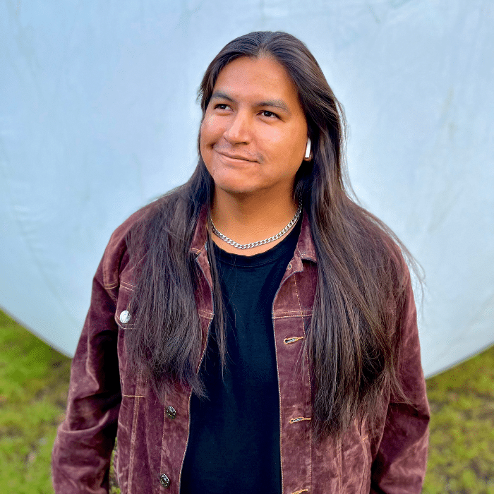A man with long dark hair stands outdoors on grass in front of a light-coloured backdrop. He is, smiling while looking slightly upward. He is wearing a maroon corduroy jacket over a black shirt, a silver chain necklace, and wireless earbuds.