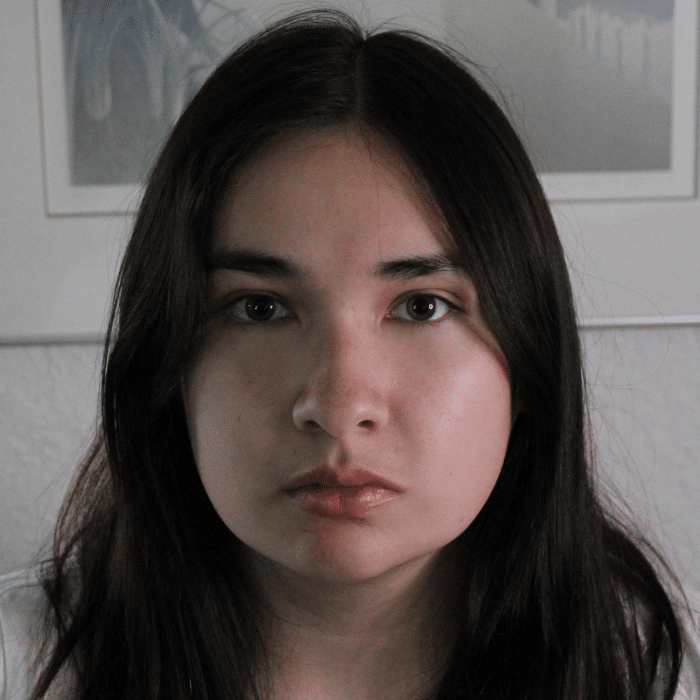 A woman with long dark hair and a neutral expression, looking directly at the camera. She is indoors with framed artwork and a textured wall in the background.