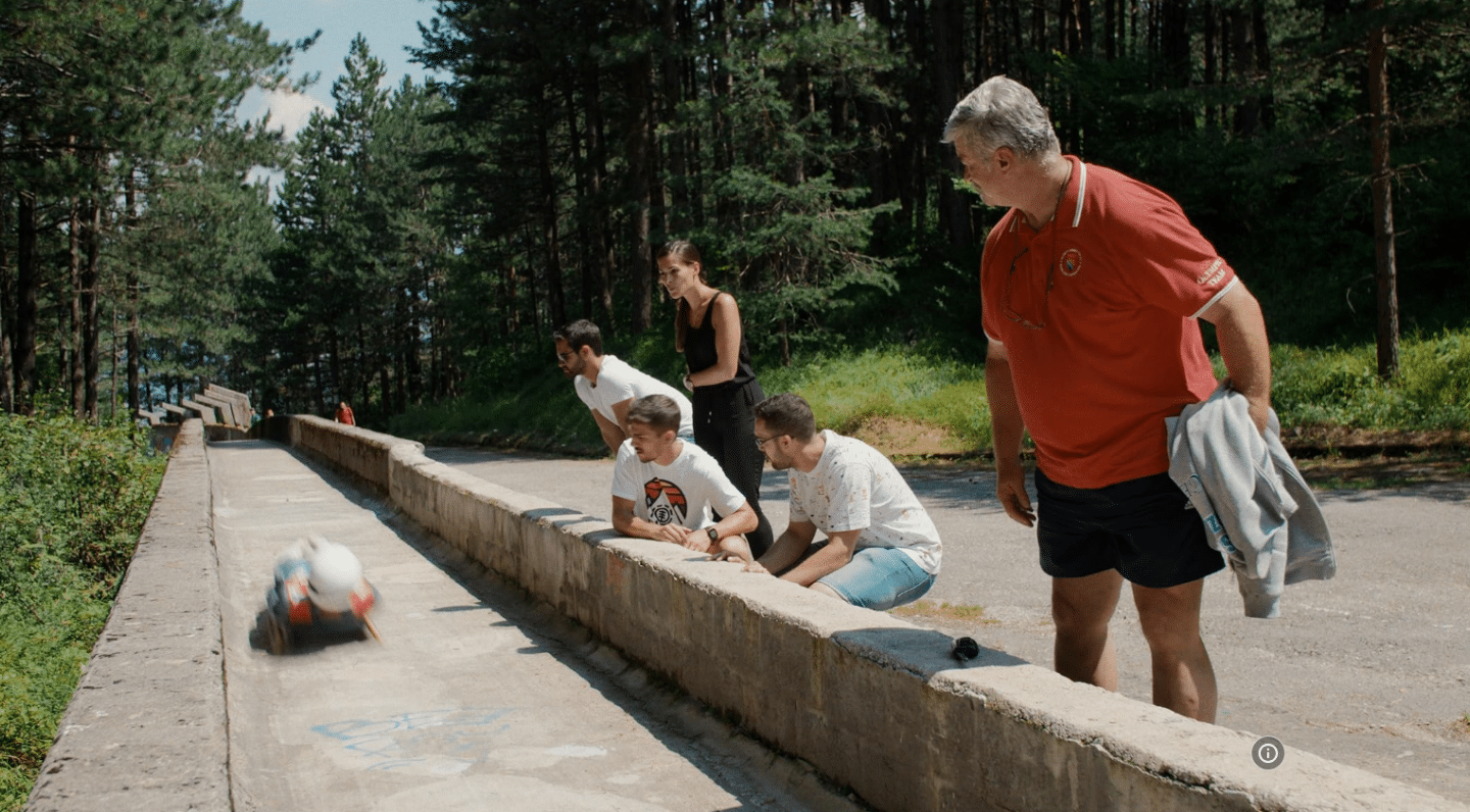 Five people lean over a short concrete wall, watching a luge and rider speed down a concrete track. There is forest on both sides of the track and the people watching.