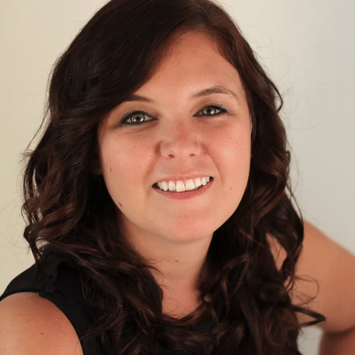 Close-up portrait of a smiling person with long, curly dark hair and light skin, wearing a black sleeveless top. She is posed against a plain light background, looking directly at the camera.
