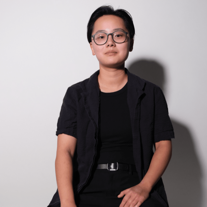 A person with short, dark hair and glasses sits slightly angled toward the camera. They wear a black shirt underneath an open short-sleeve button-up shirt against a light grey/white background.