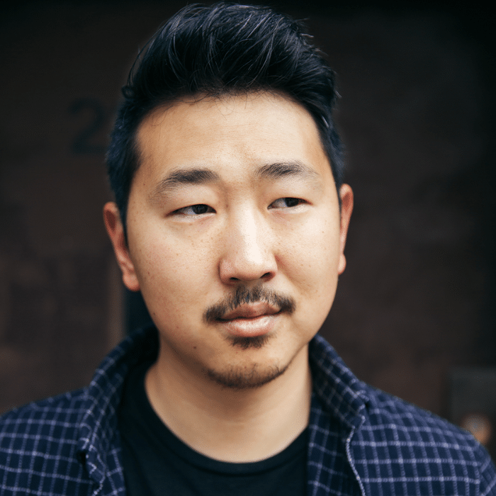 A person with short dark hair and a trimmed mustache and beard looks slightly to the side with a thoughtful expression. Andrew is wearing a black shirt and a dark checkered overshirt, photographed against a dark, softly blurred background.