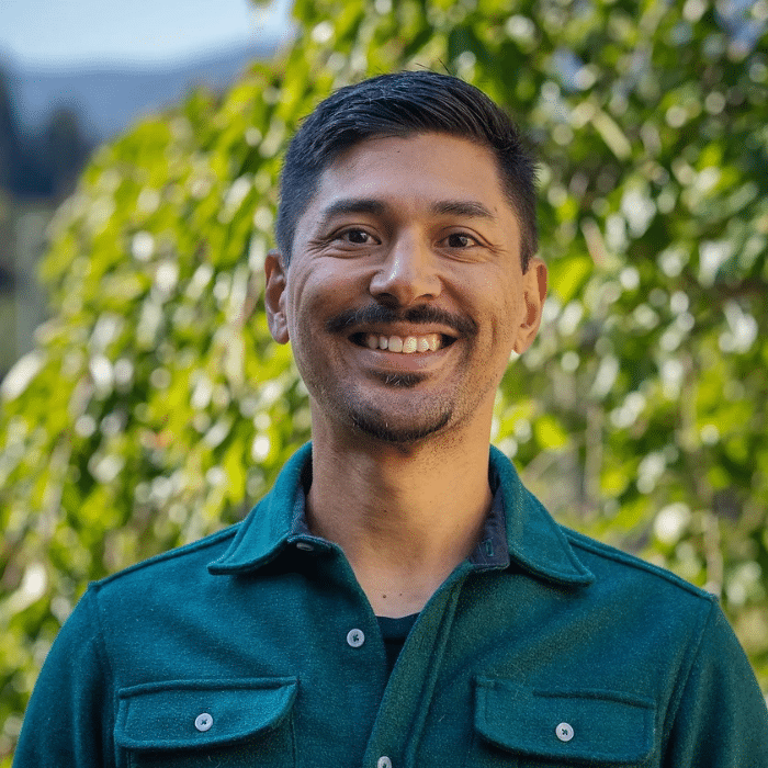 A person with short dark hair and a mustache smiles at the camera. He is wearing a teal button-up shirt, standing outdoors with bright green leaves behind him.