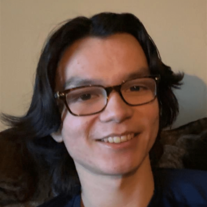 A person with medium-length dark hair and glasses smiling while sitting indoors against a beige wall. He is wearing a dark top.