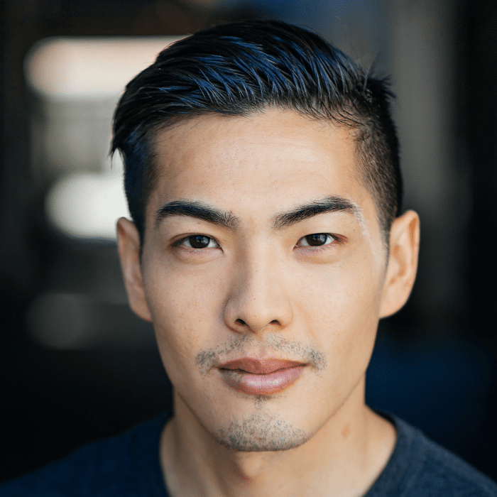A person with short dark hair and a trimmed beard looks directly at the camera with a neutral expression. He is wearing a dark shirt, and the background is blurred.