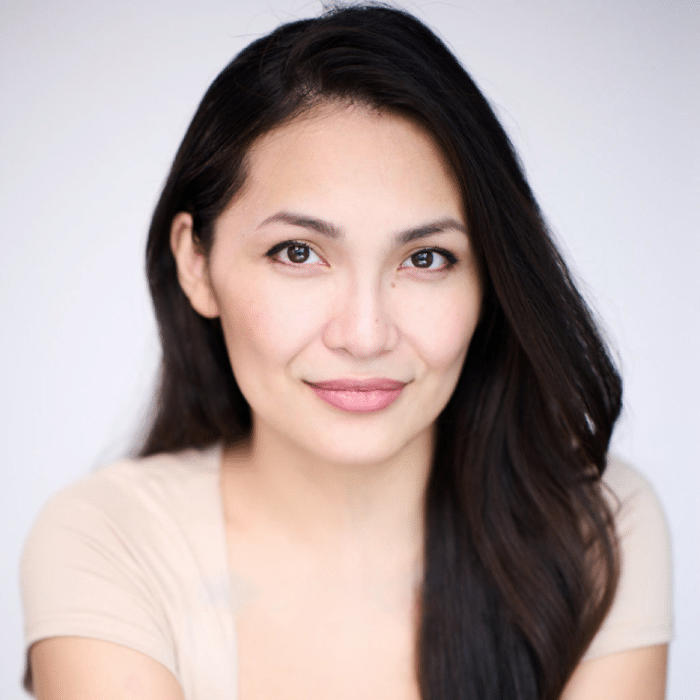 A person with long dark hair smiles at the camera. She is wearing a light-colored top, and the background is bright and plain.