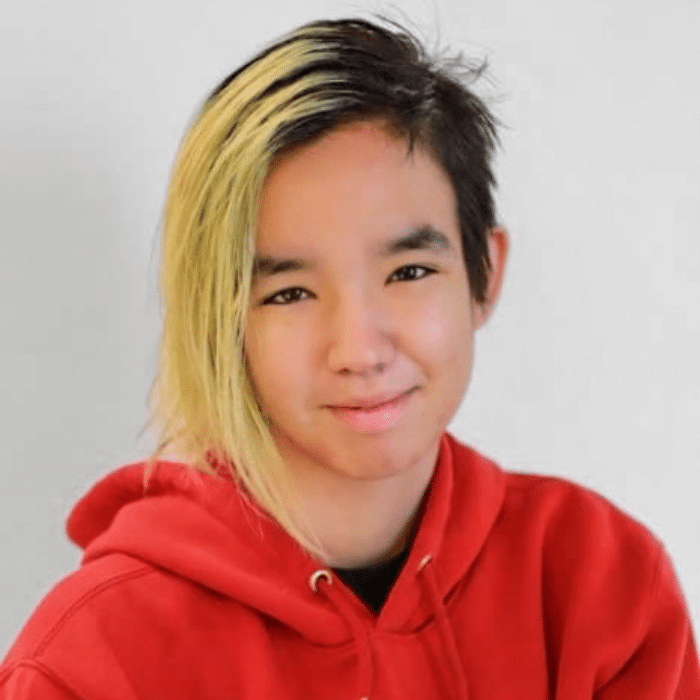 A person with short hair dyed light blonde on one side and dark on the other smiles slightly in front of a plain light background. She is wearing a bright red hoodie.