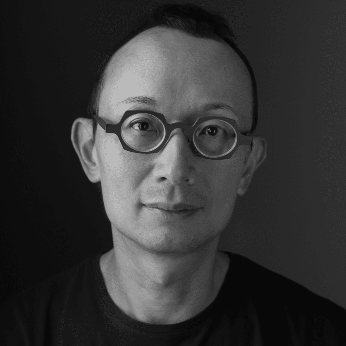 A person with short hair and distinctive geometric eyeglass frames looks directly at the camera. The portrait is black and white with a dark background.