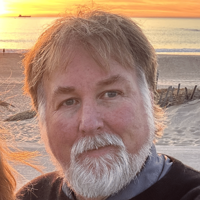 A person with sandy-coloured hair and a full white beard and moustache with a neutral expression standing on a beach at sunset. The ocean and orange sky are visible behind him.