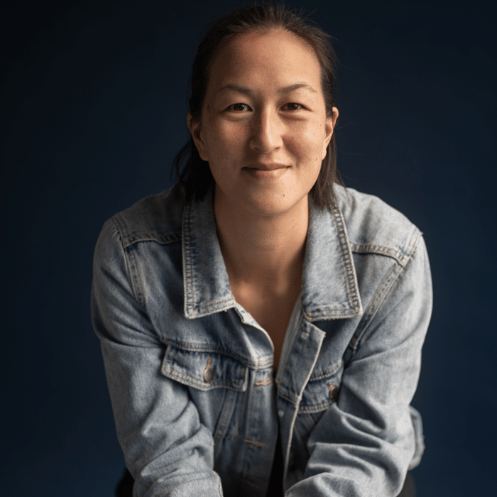 A person with dark hair pulled back. She is smiling and wearing a light-wash denim jacket and is leaning forward against a dark blue background.
