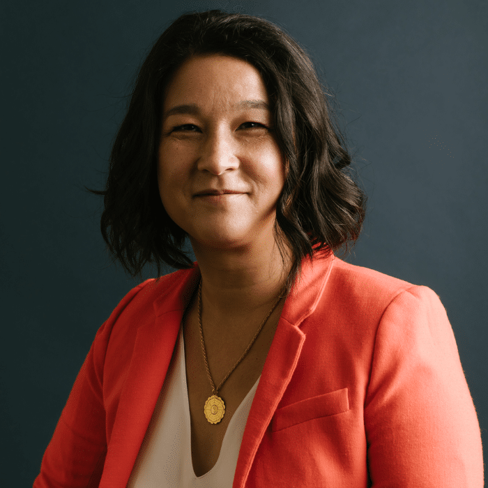A person with medium-length wavy dark hair smiling. She is wearing a coral blazer over a white blouse and a gold necklace, posed against a dark blue-grey background.