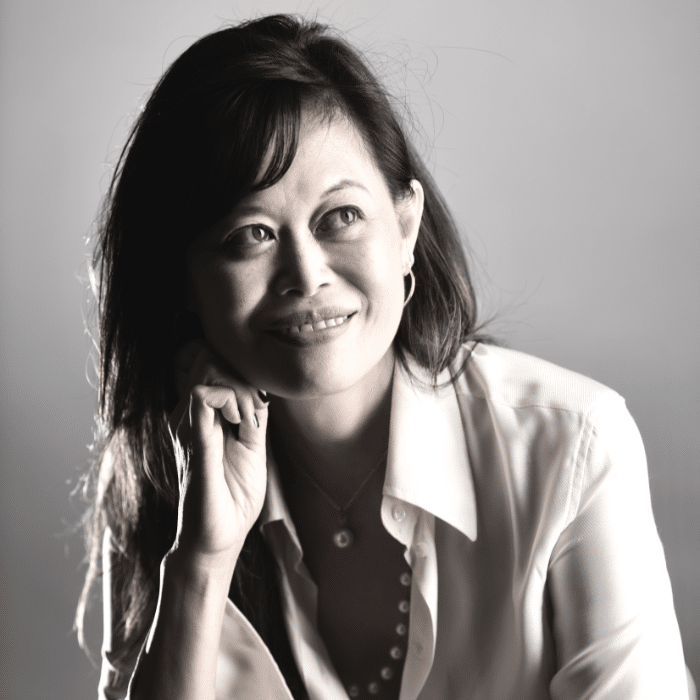 A person with long dark hair smiles while looking off to the side with a hand under her chin. She is wearing a light-colored top, and the background is bright and plain.